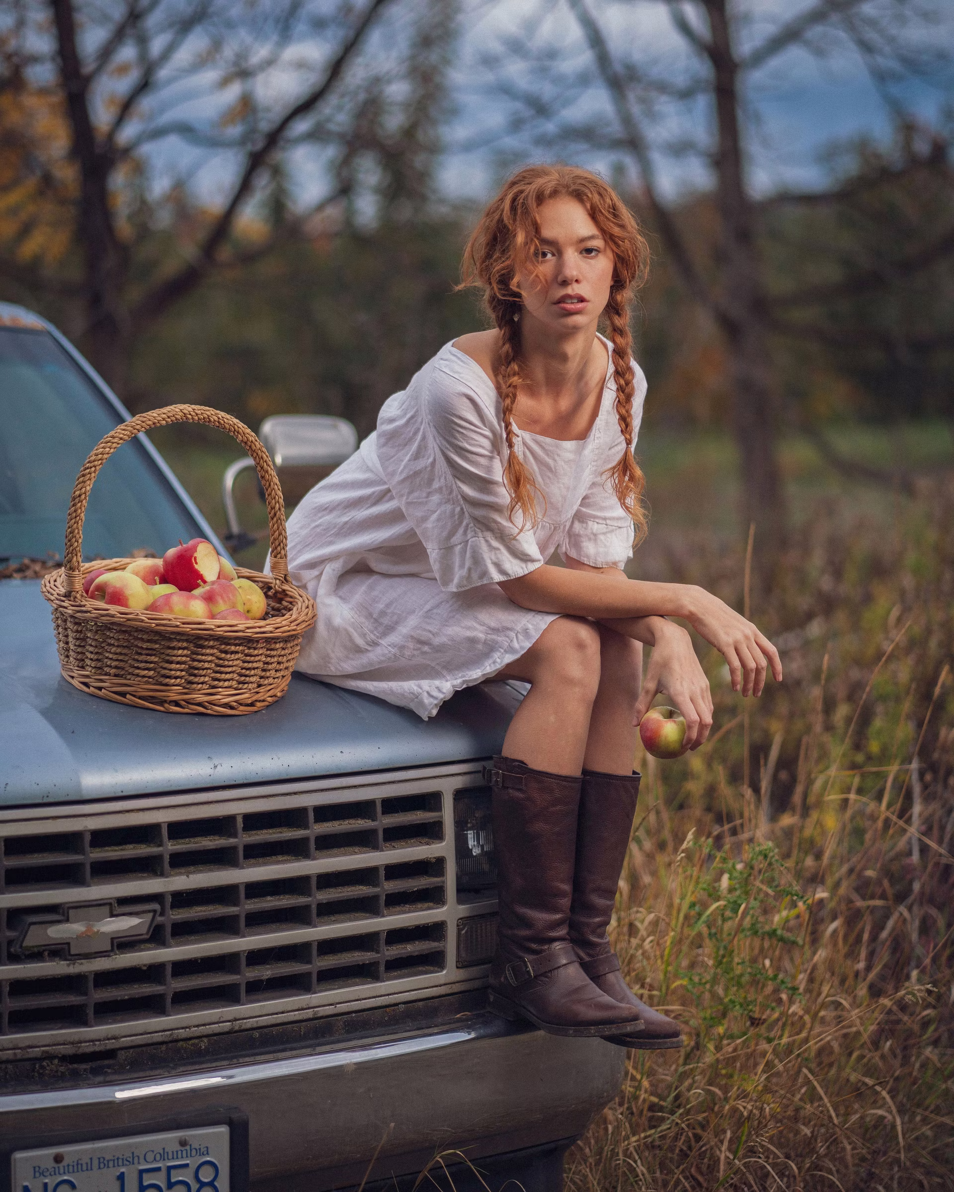 Mickie sits on the hood of a vintage blue Chevrolet pickup, white peasant dress and brown riding boots, a wicker basket of apples beside her, BC plate visible. Editorial portrait by Josiah Crowell.
