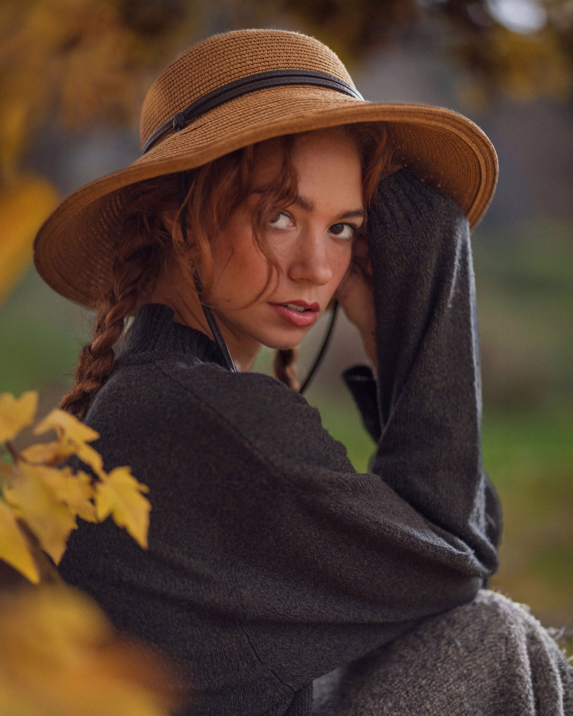 Mickie in a charcoal knit sweater and a straw boater hat, looking back over her shoulder, yellow maple leaves framing the foreground. Editorial portrait by Josiah Crowell.