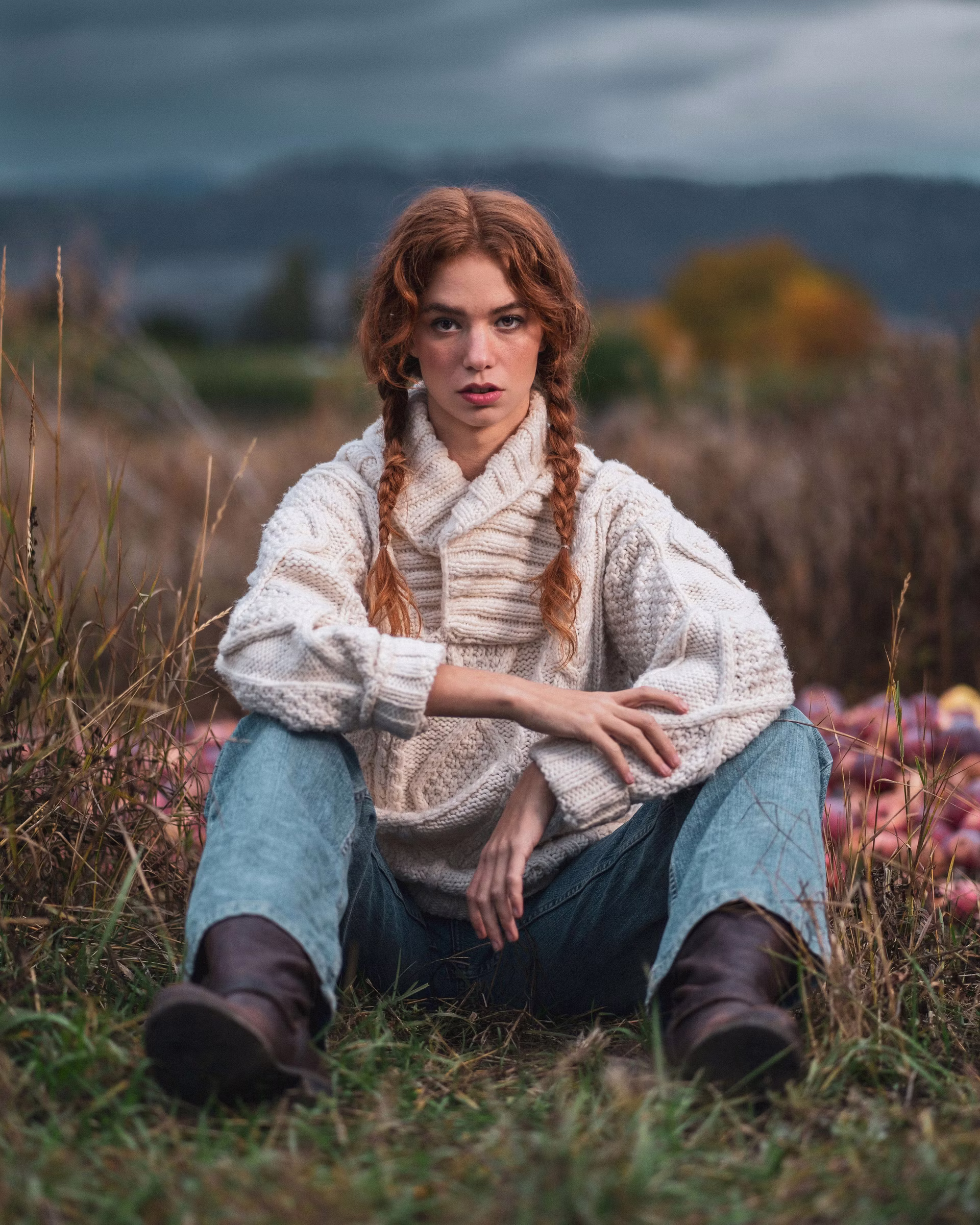 Mickie sits on the ground in tall grass wearing a cream cable-knit sweater and jeans, a pile of red apples beside her, mountains and a band of yellow trees on the horizon under a moody overcast sky. Editorial portrait by Josiah Crowell.