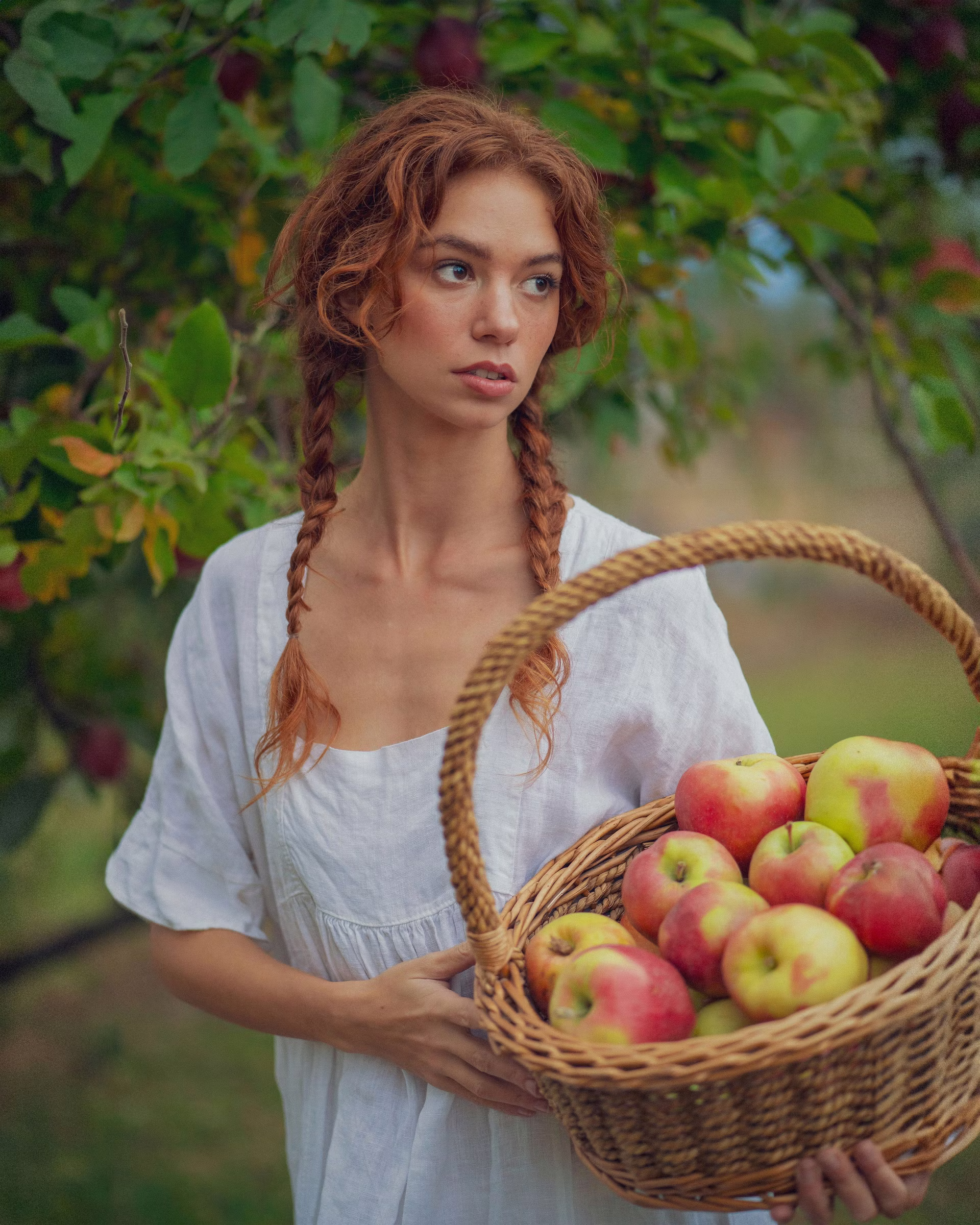 Mickie in a white peasant dress holding a wicker basket overflowing with red and green apples, standing under a tree in dappled light. Editorial portrait by Josiah Crowell.
